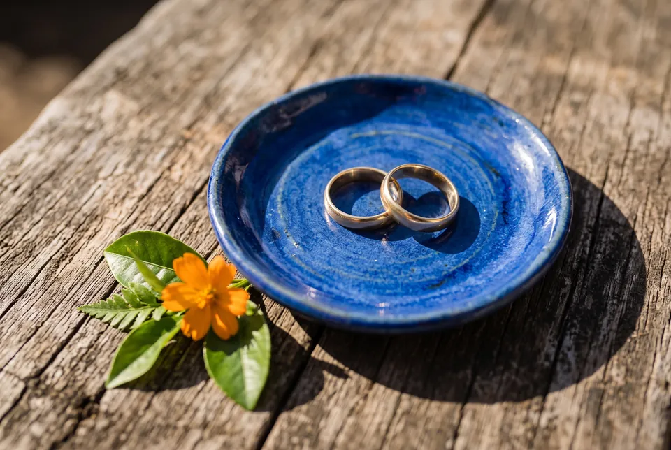 Zwei goldene Ringe in einer kobaltblauen Keramikschale, daneben eine orange Blüte
